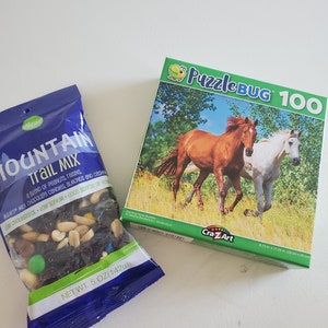 May include: A blue bag of Mountain Trail Mix with a blend of peanuts, raisins, milk chocolate candies, almonds and cashews. The bag is labeled with a net weight of 5 ounces (142g). Next to the bag is a green and white box of a 100 piece Puzzle Bug jigsaw puzzle featuring two horses running in a field. The box is labeled with the text "Puzzle Bug 100" and "Cra-Z-Art".