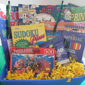 May include: A blue basket filled with yellow shredded paper and various games and activities, including a Sudoku puzzle book, a coloring book, a crossword puzzle book, a 500-piece jigsaw puzzle, a puzzle bug, and a box of colored pencils.