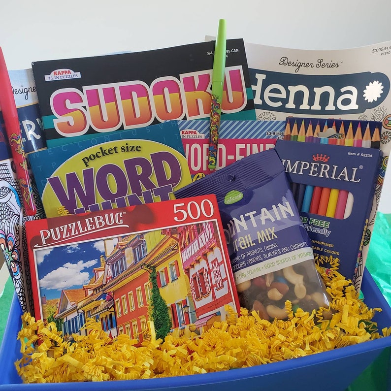 May include: A blue basket filled with a variety of items, including a 500-piece puzzle, a book of Sudoku puzzles, a book of word search puzzles, a coloring book, a box of colored pencils, and a bag of trail mix.
