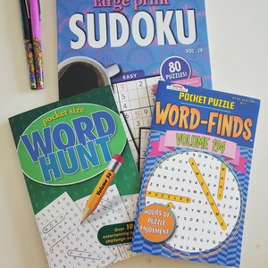 May include: Three paperback books with word puzzles. The top book is a large print Sudoku puzzle book with a blue and pink cover. The middle book is a green and white word hunt puzzle book. The bottom book is a blue and yellow word find puzzle book. All three books have the words "Pocket Puzzle" on the cover.