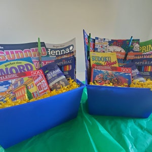 May include: Two blue baskets filled with yellow shredded paper and various items, including coloring books, pencils, and puzzles. The baskets are on a green surface. The coloring books include titles like "Sudoku Word", "Sudoku Plus", and "Henna".