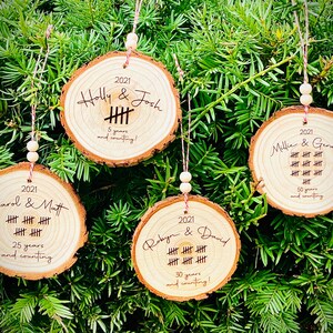 May include: Four wooden ornaments with engraved text and tally marks. The ornaments are decorated with the names of couples and the number of years they have been together. The ornaments are hanging on a green Christmas tree.