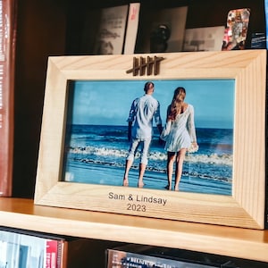 May include: A wooden picture frame with a photo of a couple on a beach. The frame has a decorative metal accent and the text "Sam & Lindsay 2023" is engraved on the bottom.