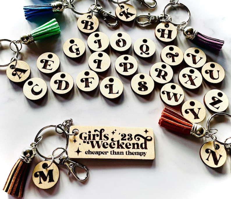 May include: A set of wooden alphabet letter keychains with brown tassels. The keychains are arranged in alphabetical order. A wooden keychain with the text "Girls '23 Weekend cheaper than therapy" is also included.