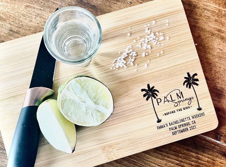 May include: A wooden cutting board with a printed design of palm trees and the text "PALM Springs -BEFORE THE RING- EMMA'S BACHELORETTE WEEKEND PALM SPRINGS, CA SEPTEMBER 2021". There is a shot glass, a lime wedge, and a knife on the board.