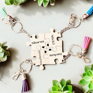 K&ouml;nnte beinhalten: Vier silberne Puzzleteile mit den Namen Sharon, Nikki, Stephanie und Darlene eingraviert. Jedes Teil hat einen kleinen Herz-Ausschnitt und ist durch eine silberne Kette mit den anderen Teilen verbunden. Die Puzzleteile sind an Schl&uuml;sselringen mit bunten Quasten befestigt.