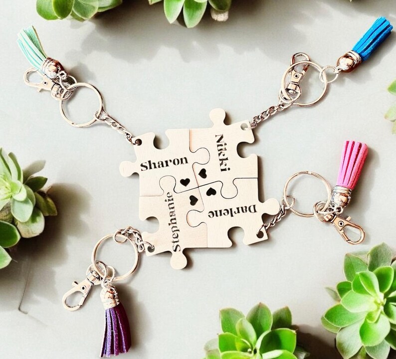 K&ouml;nnte beinhalten: Vier silberne Puzzleteile mit den Namen Sharon, Nikki, Stephanie und Darlene eingraviert. Jedes Teil hat einen kleinen Herz-Ausschnitt und ist durch eine silberne Kette mit den anderen Teilen verbunden. Die Puzzleteile sind an Schl&uuml;sselringen mit bunten Quasten befestigt.