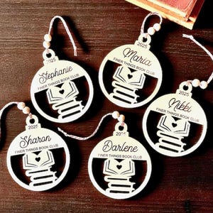 May include: Five white wooden ornaments with names and the text "FINER THINGS BOOK CLUB" and the year 2025. Each ornament features a stack of books with a heart. The names on the ornaments are Stephanie, Maria, Nikki, Sharon, and Darlene.