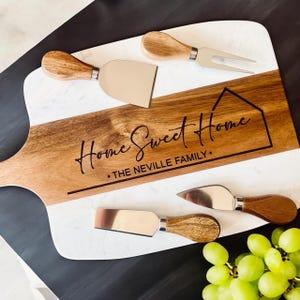 May include: A wooden and marble cheese board with four cheese knives and a fork. The board is engraved with "Home Sweet Home" and "THE NEVILLE FAMILY." The knives and fork have wooden handles and silver blades. Green grapes are in the foreground.