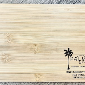 May include: A wooden cutting board with a black design of palm trees and the text "PALM Springs -BEFORE THE RING- EMMA'S BACHELORETTE WEEKEND PALM SPRINGS, CA SEPTEMBER 2021"