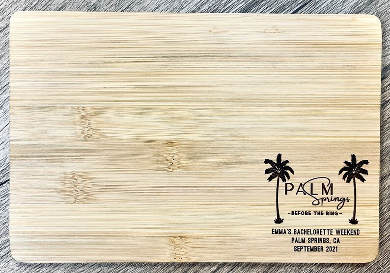 May include: A wooden cutting board with a black design of palm trees and the text "PALM Springs -BEFORE THE RING- EMMA'S BACHELORETTE WEEKEND PALM SPRINGS, CA SEPTEMBER 2021"