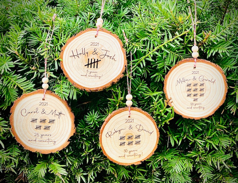 May include: Four wooden ornaments with engraved text and tally marks. The ornaments are decorated with the names of couples and the number of years they have been together. The ornaments are hung on a green Christmas tree.