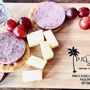 May include: A wooden cutting board with a variety of snacks, including crackers, cheese, and grapes. The board is decorated with a palm tree design and the text "PALM Springs - BEFORE THE RING - EMMA'S BACHELORETTE WEEKEND PALM SPRINGS, CA SEPTEMBER 2021".