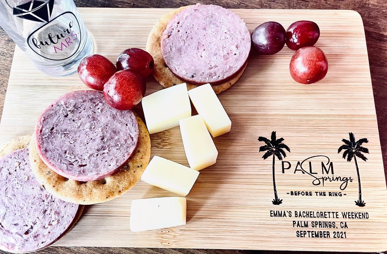 May include: A wooden cutting board with a variety of snacks, including crackers, cheese, and grapes. The board is decorated with a palm tree design and the text "PALM Springs - BEFORE THE RING - EMMA'S BACHELORETTE WEEKEND PALM SPRINGS, CA SEPTEMBER 2021".
