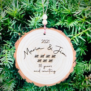 May include: A wooden round ornament with the text "2021 Maria & Jim" and a series of hash marks representing 35 years. The text "35 years and counting" is also included.