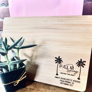 May include: A wooden cutting board with the text "Palm Springs - Before the Ring - Emma's Bachelorette Weekend Palm Springs, CA September 2021" engraved on it. Two palm trees are also engraved on the board.
