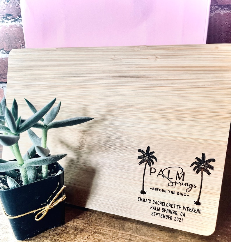 May include: A wooden cutting board with the text "Palm Springs - Before the Ring - Emma's Bachelorette Weekend Palm Springs, CA September 2021" engraved on it. Two palm trees are also engraved on the board.