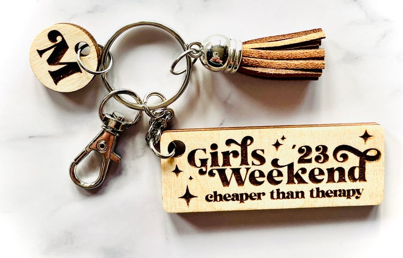 May include: Wooden keychain with a brown leather tassel, a silver metal ring, and a wooden charm with the text "Girls '23 Weekend cheaper than therapy".