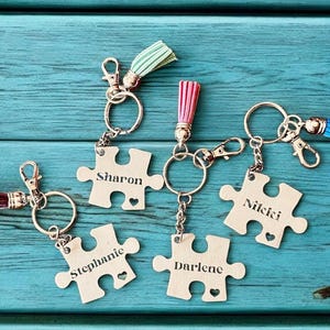 K&ouml;nnte beinhalten: F&uuml;nf silberne Puzzle-Schl&uuml;sselanh&auml;nger mit verschiedenen Namen, die darauf graviert sind. Jeder Schl&uuml;sselanh&auml;nger hat einen kleinen Quastenanh&auml;nger. Die Namen auf den Schl&uuml;sselanh&auml;ngern sind Sharon, Stephanie, Darlene, Nicki und ein f&uuml;nfter Schl&uuml;sselanh&auml;nger mit einer blauen Quaste.