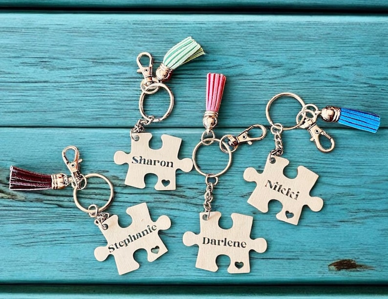 K&ouml;nnte beinhalten: F&uuml;nf silberne Puzzle-Schl&uuml;sselanh&auml;nger mit verschiedenen Namen, die darauf graviert sind. Jeder Schl&uuml;sselanh&auml;nger hat einen kleinen Quastenanh&auml;nger. Die Namen auf den Schl&uuml;sselanh&auml;ngern sind Sharon, Stephanie, Darlene, Nicki und ein f&uuml;nfter Schl&uuml;sselanh&auml;nger mit einer blauen Quaste.