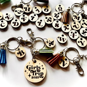 May include: A set of wooden keychains with letters of the alphabet, a keychain with the words "Girls Trip 2023" and a keychain with the letter "N". Each keychain has a small tassel attached.