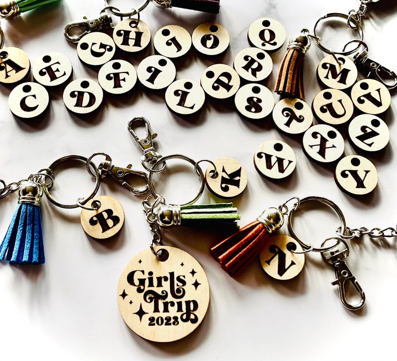 May include: A set of wooden keychains with letters of the alphabet, a keychain with the words "Girls Trip 2023" and a keychain with the letter "N". Each keychain has a small tassel attached.