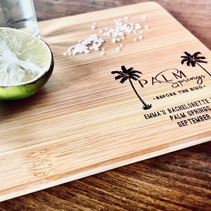May include: A wooden cutting board with the text "Palm Springs - Before the Ring" and "Emma's Bachelorette Weekend Palm Springs, CA September 2021". There are two palm trees and a lime wedge on the board.