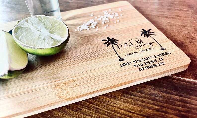 May include: A wooden cutting board with the text "Palm Springs - Before the Ring" and "Emma's Bachelorette Weekend Palm Springs, CA September 2021". There are two palm trees and a lime wedge on the board.