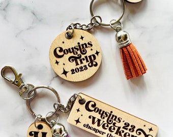 Könnte beinhalten: Zwei Holzschlüsselanhänger mit Quasten. Ein Schlüsselanhänger hat ein rundes Schild mit dem Text "Cousins Trip 2023" und der andere Schlüsselanhänger hat ein rechteckiges Schild mit dem Text "Cousins '23 Weekend cheaper than therapy".