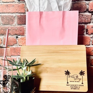 May include: A wooden cutting board with a laser engraved design featuring palm trees and the text "Palm Springs - Before the Ring - Emma's Bachelorette Weekend Palm Springs, CA September 2021".