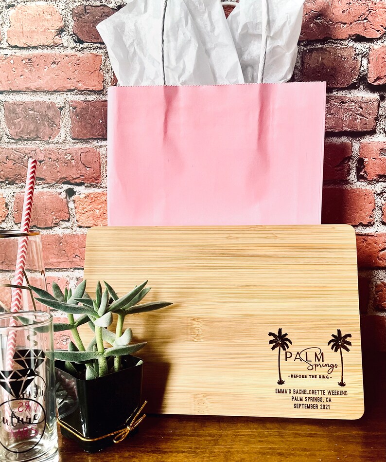 May include: A wooden cutting board with a laser engraved design featuring palm trees and the text "Palm Springs - Before the Ring - Emma's Bachelorette Weekend Palm Springs, CA September 2021".