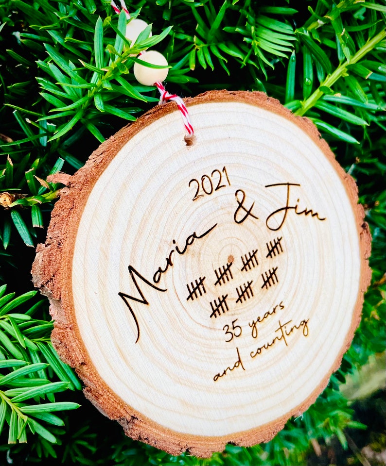 May include: A wooden ornament with the text "2021 Maria & Jim" and "35 years and counting" engraved on it. The ornament is decorated with hash marks to represent the years.
