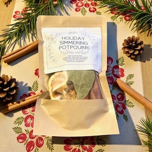 May include: A kraft paper bag containing holiday simmering potpourri. The bag has a clear window showing dried lemon slices, bay leaves, and spices. The label reads "Holiday Simmering Potpourri". Cinnamon sticks, pine needles, and a pine cone are arranged around the bag.