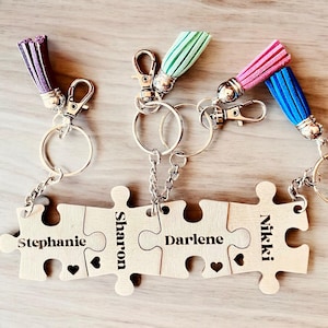 K&ouml;nnte beinhalten: Vier Holzteile eines Puzzles mit den Namen Stephanie, Sharon, Darlene und Nikki eingraviert. Die Teile sind durch Schl&uuml;sselanh&auml;nger mit bunten Quasten verbunden.