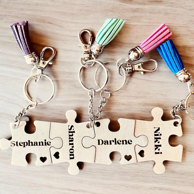 K&ouml;nnte beinhalten: Vier Holzteile eines Puzzles mit den Namen Stephanie, Sharon, Darlene und Nikki eingraviert. Die Teile sind durch Schl&uuml;sselanh&auml;nger mit bunten Quasten verbunden.