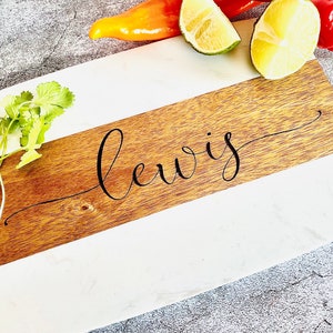 May include: A white and brown marble cutting board with the name "Lewis" engraved on the wood portion. The board is decorated with fresh cilantro and limes.