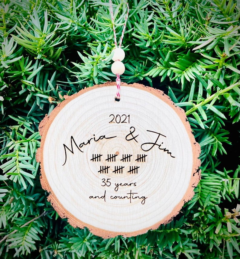May include: A wooden round ornament with the text "2021 Maria & Jim" and a series of hash marks representing 35 years. The text "35 years and counting" is also included.