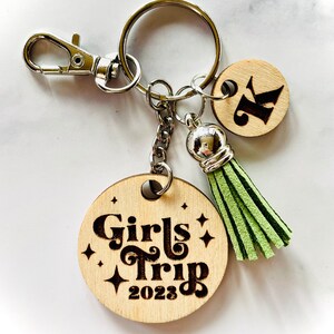 May include: A wooden keychain with a green tassel and a round wooden charm that says "Girls Trip 2023" in a retro font. There is also a smaller round wooden charm with the letter "K" on it.