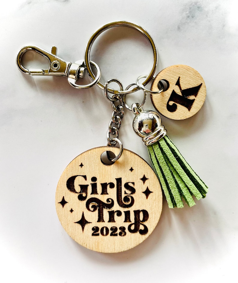 May include: A wooden keychain with a green tassel and a round wooden charm that says "Girls Trip 2023" in a retro font. There is also a smaller round wooden charm with the letter "K" on it.