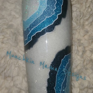May include: A white tumbler with a blue and black agate design. The tumbler has a glittery finish and the text "Munchkin Mama Designs" is written on the side.