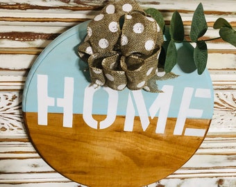 Può includere: Un cartello rotondo in legno con la parola "HOME" dipinta in bianco su uno sfondo blu chiaro. Il cartello è decorato con un fiocco a pois marroni e bianchi e un rametto di eucalipto.