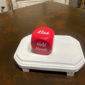 May include: Red dice with white lettering. The dice has the words "Kiss" and "Hold Hands" on it. The dice is sitting on a white pedestal.