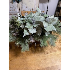 Puede incluir: Un centro de mesa con poinsettias verdes escarchadas, ramas de pino y piñas en una cesta tejida.