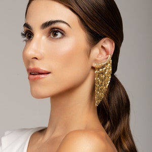 Puede incluir: Una mujer con el pelo largo y castaño lleva un pendiente de cadena dorada en forma de aro. El pendiente es de estilo largo y colgante con una franja de pequeñas cadenas doradas.