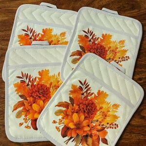 May include: Set of six white quilted pot holders with a floral design featuring orange, yellow, and brown leaves and flowers.
