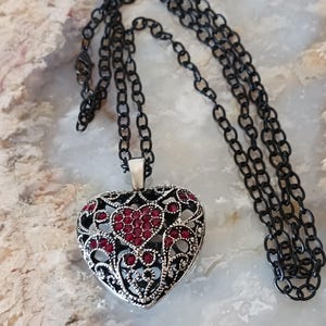 May include: A heart-shaped pendant necklace with a black chain. The pendant is silver-toned with an open filigree design and is adorned with red gemstones. The necklace is displayed on a light-coloured, marbled surface.