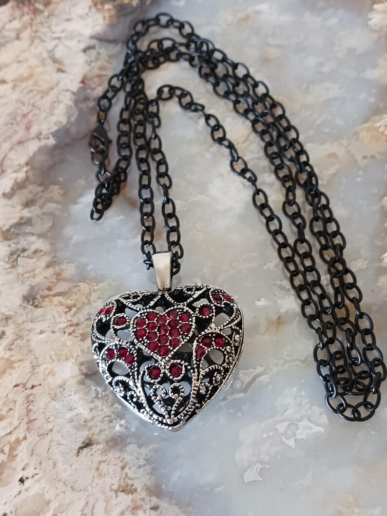 May include: A heart-shaped pendant necklace with a black chain. The pendant is silver-toned with an open filigree design and is adorned with red gemstones. The necklace is displayed on a light-coloured, marbled surface.