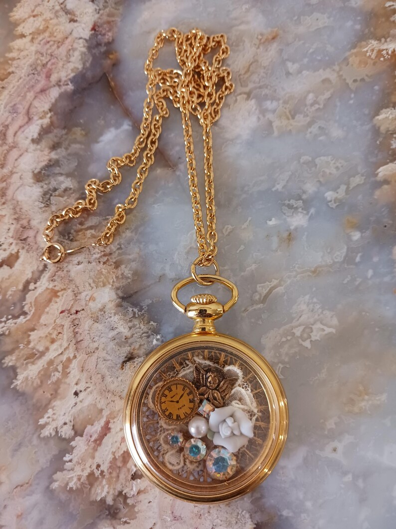 Puede incluir: Collar con colgante dorado con un medall&oacute;n redondo y transparente. Dentro del medall&oacute;n hay elementos decorativos en miniatura: un peque&ntilde;o reloj, una figura de &aacute;ngel, perlas y cristales. El collar tiene una cadena fina.