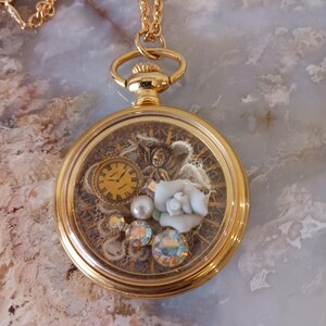Puede incluir: Collar con colgante tipo relicario dorado. Presenta una esfera transparente que revela una escena en miniatura. Contiene un peque&ntilde;o reloj, una rosa blanca, perlas y gemas iridiscentes. La cadena tambi&eacute;n es dorada.