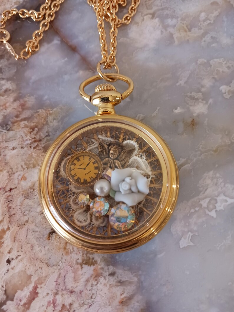 Puede incluir: Collar con colgante tipo relicario dorado. Presenta una esfera transparente que revela una escena en miniatura. Contiene un peque&ntilde;o reloj, una rosa blanca, perlas y gemas iridiscentes. La cadena tambi&eacute;n es dorada.